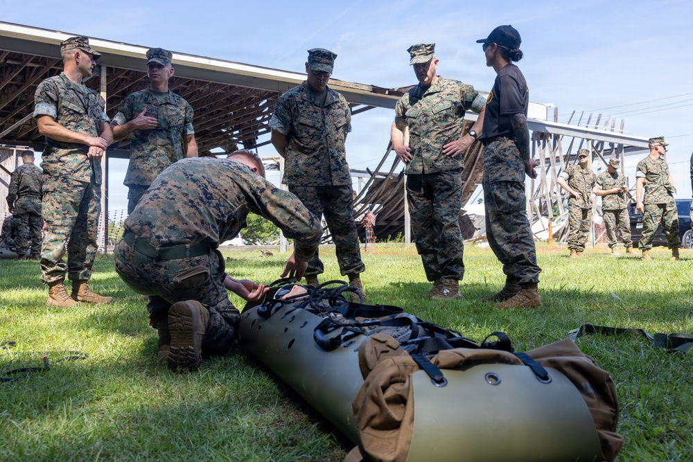 A group of military personnel in MARPAT camouflage uniforms, consistent with U.S. Marines, are gathered outdoors on a grass field near a damaged or partially collapsed structure. One service member is crouched down handling what appears to be a military litter or rescue stretcher on the ground. The scene appears to be a training exercise or post-disaster assessment activity.
