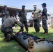 A group of military personnel in MARPAT camouflage uniforms, consistent with U.S. Marines, are gathered outdoors on a grass field near a damaged or partially collapsed structure. One service member is crouched down handling what appears to be a military litter or rescue stretcher on the ground. The scene appears to be a training exercise or post-disaster assessment activity.
