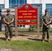 Four U.S. Marines in woodland MARPAT camouflage uniforms stand in front of an official unit sign for the Expeditionary Operations Training Group, II Marine Expeditionary Force. The sign identifies the OIC as Col N.R. Berry and the SNCOIC as MGySgt R.G. Smith. This appears to be a formal unit photo taken at a military installation.