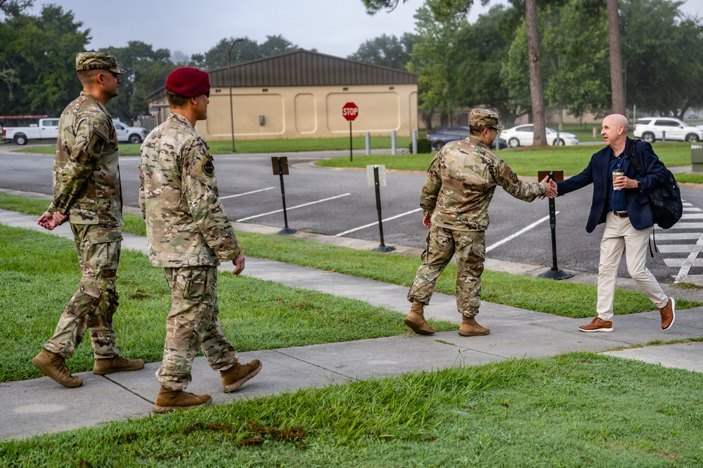 Three military personnel in camouflage uniforms are walking along a sidewalk on what appears to be a military installation. A civilian man in a navy blazer and khaki pants is approaching and extending his hand toward one of the soldiers in a greeting gesture. The setting includes a parking lot, a stop sign, a tan building, and well-maintained grass.