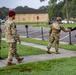 Three military personnel in camouflage uniforms are walking along a sidewalk on what appears to be a military installation. A civilian man in a navy blazer and khaki pants is approaching and extending his hand toward one of the soldiers in a greeting gesture. The setting includes a parking lot, a stop sign, a tan building, and well-maintained grass.