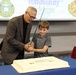 An older man and a young boy are together cutting a large sheet cake using a ceremonial sword. The setting appears to be a formal military or community event, evidenced by the insignia and Latin motto 'Pro Deo Et Patria' visible on the projection screen behind them. The boy is smiling, suggesting this is a celebratory occasion.