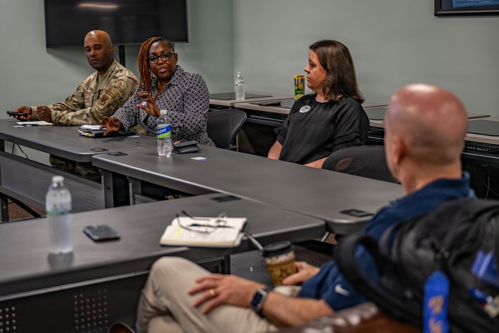 A small group meeting or briefing is taking place in a conference or classroom setting. Four individuals are visible: one person in military camouflage uniform, two women seated at a table, and one bald man seated in the foreground with his back to the camera. The woman in the center appears to be speaking or presenting to the group.