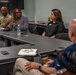 A small group meeting or briefing is taking place in a conference or classroom setting. Four individuals are visible: one person in military camouflage uniform, two women seated at a table, and one bald man seated in the foreground with his back to the camera. The woman in the center appears to be speaking or presenting to the group.