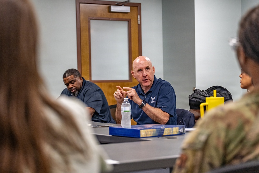 A bald man wearing a navy blue polo shirt with a U.S. Air Force logo is speaking and gesturing with his hands at a conference table. He appears to be leading or participating in a briefing or discussion with several other individuals, including a person in military camouflage uniform visible in the foreground. A water bottle and blue book/binder are on the table in front of him.