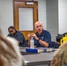 A bald man wearing a navy blue polo shirt with a U.S. Air Force logo is speaking and gesturing with his hands at a conference table. He appears to be leading or participating in a briefing or discussion with several other individuals, including a person in military camouflage uniform visible in the foreground. A water bottle and blue book/binder are on the table in front of him.
