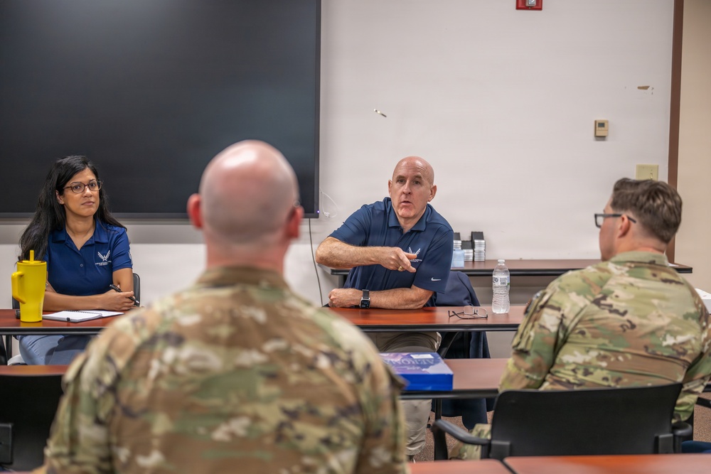 A meeting or briefing is taking place in what appears to be a classroom or conference room setting. Two civilians in blue polo shirts bearing a military insignia are seated at a table facing two uniformed military personnel in camouflage (OCP/MultiCam) uniforms. The male civilian appears to be actively speaking and gesturing toward the military personnel.