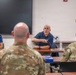 A meeting or briefing is taking place in what appears to be a classroom or conference room setting. Two civilians in blue polo shirts bearing a military insignia are seated at a table facing two uniformed military personnel in camouflage (OCP/MultiCam) uniforms. The male civilian appears to be actively speaking and gesturing toward the military personnel.