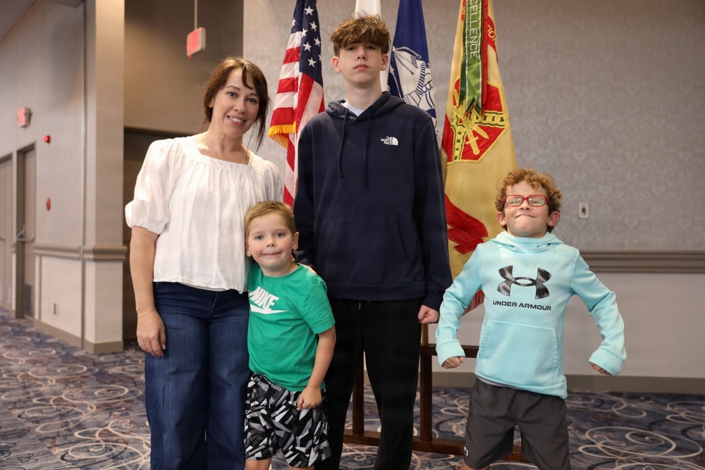 A woman and three children pose for a photo in what appears to be a military or government building. They are standing in front of several flags, including the American flag and what appears to be U.S. Army flags. The group appears to be attending an official ceremony or event.