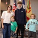 A woman and three children pose for a photo in what appears to be a military or government building. They are standing in front of several flags, including the American flag and what appears to be U.S. Army flags. The group appears to be attending an official ceremony or event.