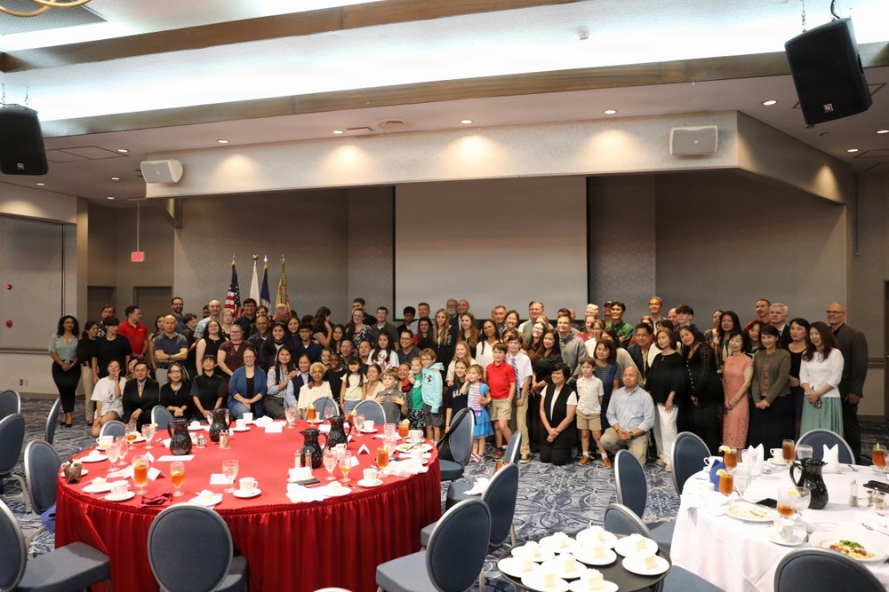 A large group of approximately 80-100 people, including adults and children, are gathered for a formal banquet or celebratory event in a hotel ballroom or conference center. The group is posed together for a group photograph in front of projection screens and what appear to be flags, including a U.S. flag. The setting features round tables with red and white tablecloths, place settings, and beverages.