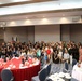 A large group of approximately 80-100 people, including adults and children, are gathered for a formal banquet or celebratory event in a hotel ballroom or conference center. The group is posed together for a group photograph in front of projection screens and what appear to be flags, including a U.S. flag. The setting features round tables with red and white tablecloths, place settings, and beverages.
