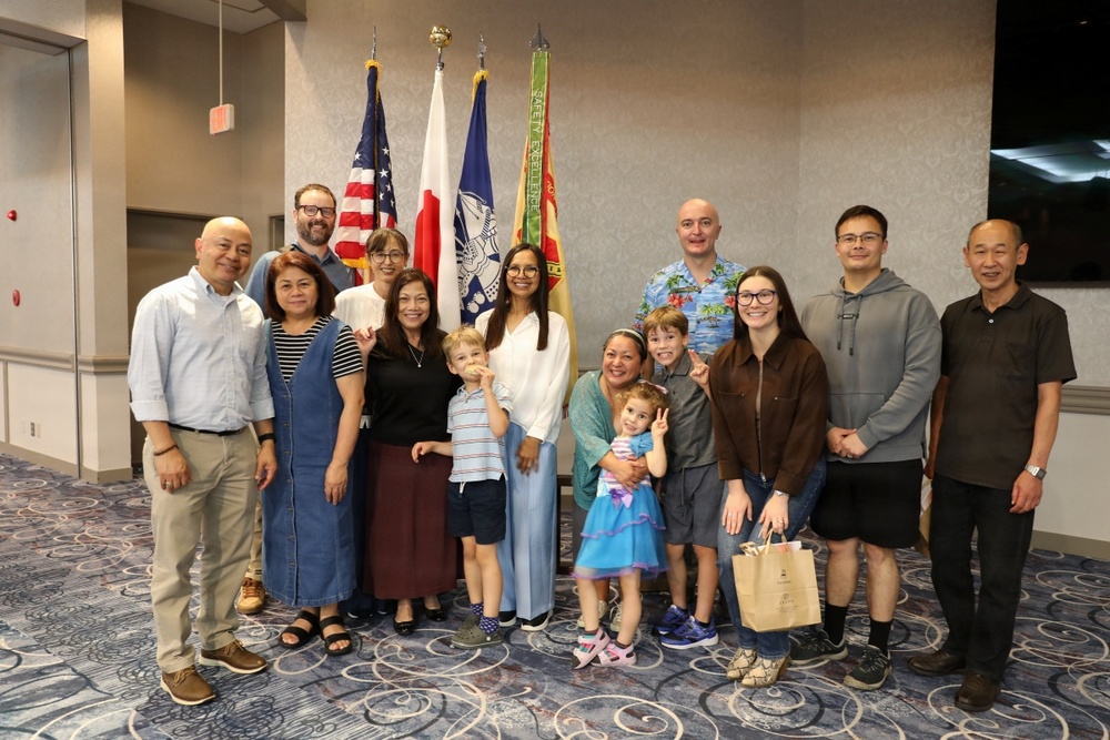 A group of approximately 14 people, including adults and young children, pose together for a formal group photo in what appears to be a government or military facility. Several flags are visible in the background, including the United States flag, a Japanese flag, a U.S. Army flag, and what appears to be a safety/excellence banner. The gathering appears to be a celebratory or official event.