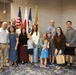 A group of approximately 14 people, including adults and young children, pose together for a formal group photo in what appears to be a government or military facility. Several flags are visible in the background, including the United States flag, a Japanese flag, a U.S. Army flag, and what appears to be a safety/excellence banner. The gathering appears to be a celebratory or official event.
