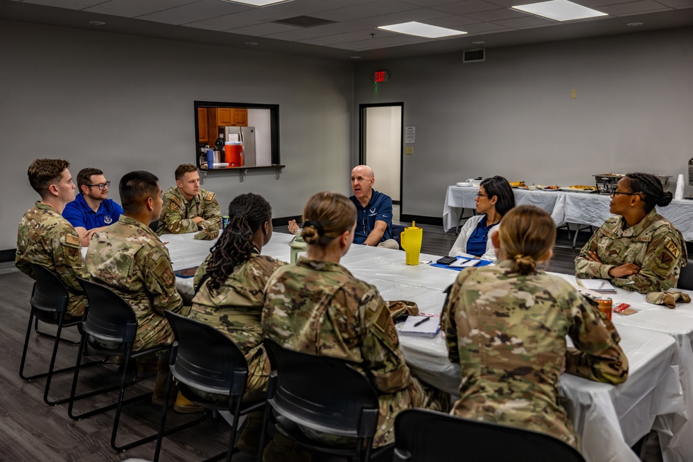 A group of approximately 11 military personnel in OCP (Operational Camouflage Pattern) uniforms are seated around a large round table in a conference or meeting room. Two civilians in blue polo shirts, one of whom appears to be speaking, are engaged in what appears to be a briefing or discussion session. A food/refreshment table with white tablecloths is visible in the background.
