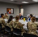 A group of approximately 11 military personnel in OCP (Operational Camouflage Pattern) uniforms are seated around a large round table in a conference or meeting room. Two civilians in blue polo shirts, one of whom appears to be speaking, are engaged in what appears to be a briefing or discussion session. A food/refreshment table with white tablecloths is visible in the background.