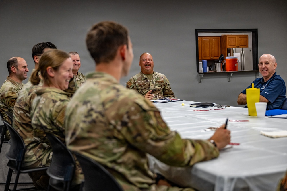 A group of military personnel in camouflage uniforms are seated around a white-cloth-covered table in what appears to be a briefing or meeting room. The atmosphere is relaxed and jovial, with several individuals visibly laughing. One civilian in a blue polo shirt is also present at the table.