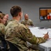 A group of military personnel in camouflage uniforms are seated around a white-cloth-covered table in what appears to be a briefing or meeting room. The atmosphere is relaxed and jovial, with several individuals visibly laughing. One civilian in a blue polo shirt is also present at the table.