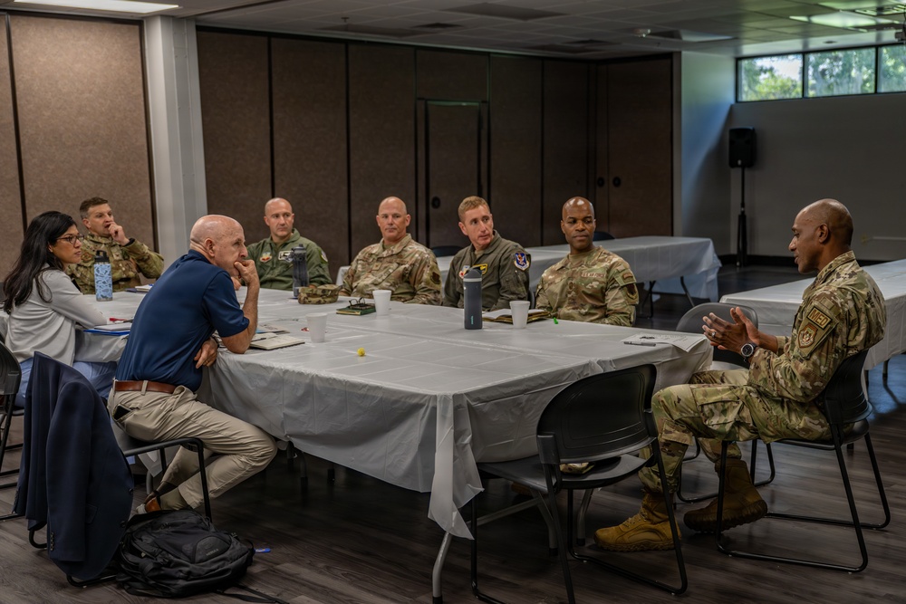 A group of approximately eight to nine individuals are seated around a rectangular table covered with a white tablecloth in what appears to be a conference or meeting room. The majority of participants are wearing U.S. military uniforms in OCP (Operational Camouflage Pattern) and flight suits, with one civilian male and one civilian female also present. A uniformed service member on the right side of the table appears to be actively speaking or briefing the group.