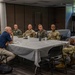 A group of approximately eight to nine individuals are seated around a rectangular table covered with a white tablecloth in what appears to be a conference or meeting room. The majority of participants are wearing U.S. military uniforms in OCP (Operational Camouflage Pattern) and flight suits, with one civilian male and one civilian female also present. A uniformed service member on the right side of the table appears to be actively speaking or briefing the group.