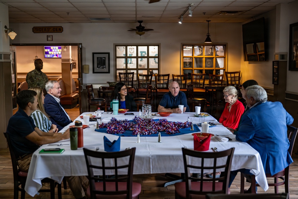 A group of approximately eight people are seated around a large rectangular table with a white tablecloth in a dining room labeled 'Tigers' Den.' The gathering appears to be a formal or semi-formal meeting or luncheon, with participants engaged in conversation. A person in military camouflage uniform is visible standing in the background near a doorway.