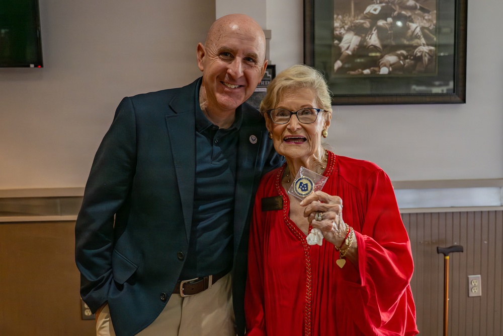 Two individuals pose together for a photo in what appears to be an indoor venue. An elderly woman in a red outfit holds up a commemorative coin or medallion in a clear plastic sleeve, suggesting a recognition or award ceremony. A framed black-and-white sports photograph is visible on the wall behind them.