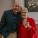 Two individuals pose together for a photo in what appears to be an indoor venue. An elderly woman in a red outfit holds up a commemorative coin or medallion in a clear plastic sleeve, suggesting a recognition or award ceremony. A framed black-and-white sports photograph is visible on the wall behind them.