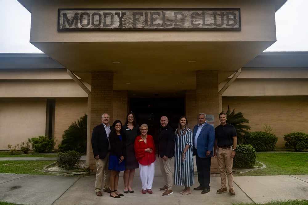 A group of eight people pose for a formal group photograph in front of the Moody Field Club building. The building features a prominent sign reading 'MOODY FIELD CLUB' above a covered entrance portico. The group appears to be gathered for an official or commemorative occasion.