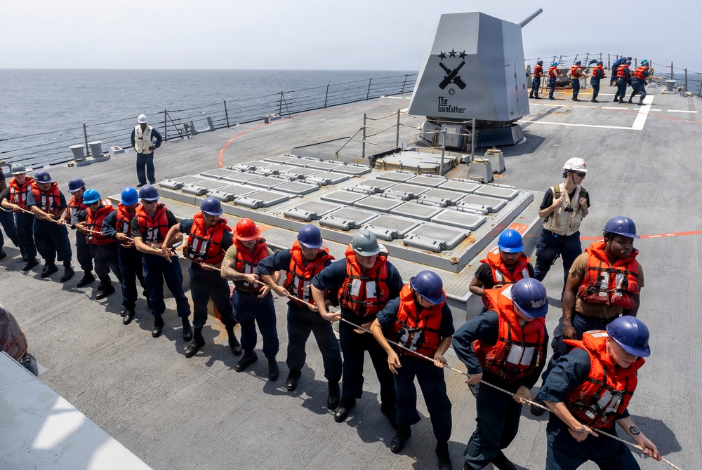 A group of U.S. Navy sailors aboard a warship are engaged in a line-handling evolution on the main deck, pulling a thick rope in unison. The sailors are wearing orange life vests and blue or red hard hats, indicating a formal seamanship drill or replenishment-at-sea operation. A naval gun mount labeled 'The Gunfather' with a crossed-rifles insignia and three stars is visible in the background, along with a Vertical Launch System (VLS) missile battery.
