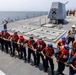 A group of U.S. Navy sailors aboard a warship are engaged in a line-handling evolution on the main deck, pulling a thick rope in unison. The sailors are wearing orange life vests and blue or red hard hats, indicating a formal seamanship drill or replenishment-at-sea operation. A naval gun mount labeled 'The Gunfather' with a crossed-rifles insignia and three stars is visible in the background, along with a Vertical Launch System (VLS) missile battery.