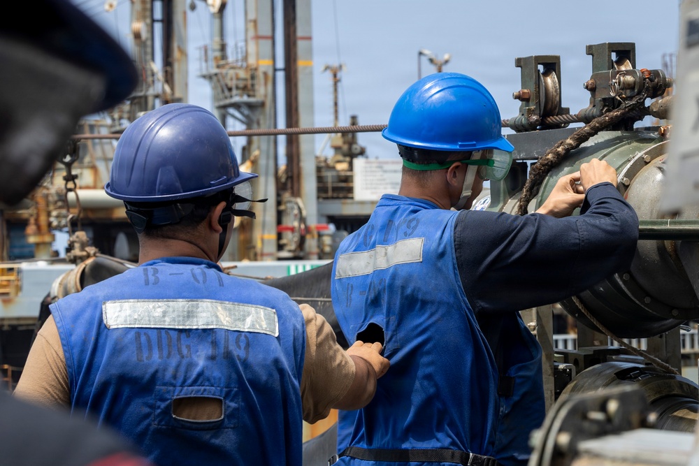 Two workers wearing blue hard hats and blue reflective safety vests marked 'DDG-119' are performing maintenance or operational tasks on heavy industrial equipment, likely aboard a naval vessel. The worker on the right is wearing additional eye protection (green safety goggles) and is manually handling a chain or rope mechanism on a large cylindrical component. The background shows a complex industrial or shipyard environment with metal structures, rigging, and equipment.