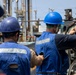 Two workers wearing blue hard hats and blue reflective safety vests marked 'DDG-119' are performing maintenance or operational tasks on heavy industrial equipment, likely aboard a naval vessel. The worker on the right is wearing additional eye protection (green safety goggles) and is manually handling a chain or rope mechanism on a large cylindrical component. The background shows a complex industrial or shipyard environment with metal structures, rigging, and equipment.