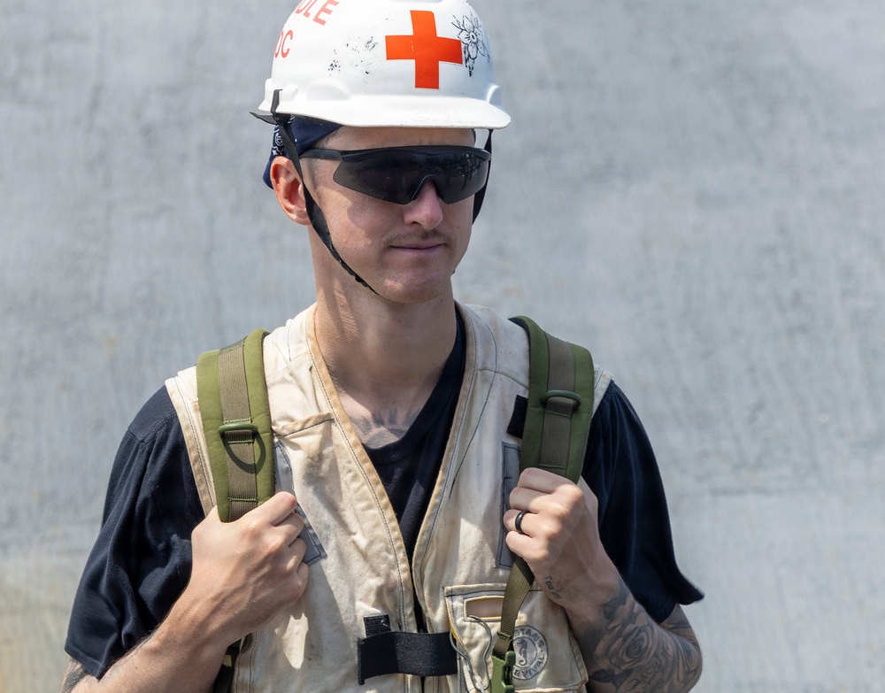 A young male wearing a white hard hat marked with a red cross symbol, dark sunglasses, and a worn tactical/utility vest over a black t-shirt. He is carrying a green backpack and has visible tattoos on his forearms. His attire suggests a role in emergency medical services, disaster relief, or a military medic context.