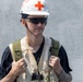 A young male wearing a white hard hat marked with a red cross symbol, dark sunglasses, and a worn tactical/utility vest over a black t-shirt. He is carrying a green backpack and has visible tattoos on his forearms. His attire suggests a role in emergency medical services, disaster relief, or a military medic context.
