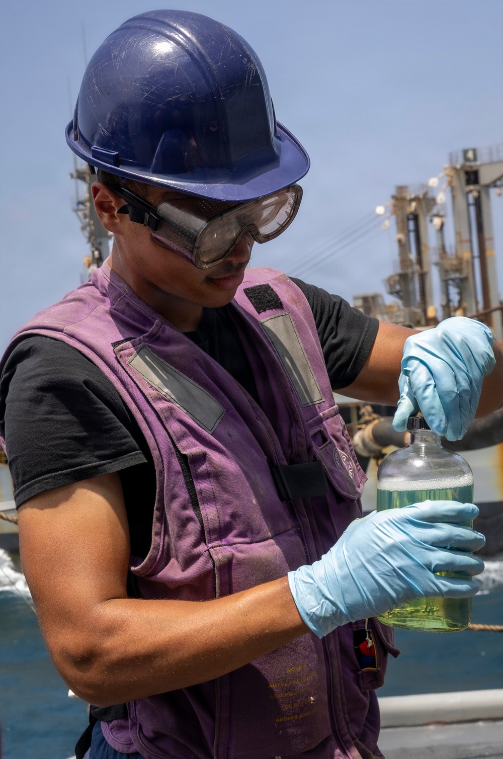 A worker wearing a blue hard hat, safety goggles, blue nitrile gloves, and a purple vest is handling a clear glass bottle containing a green/yellow liquid. The setting appears to be an outdoor industrial or maritime environment, with what looks like a naval vessel or industrial structure visible in the background. The activity suggests fuel or liquid sampling, consistent with maritime or military fueling operations.