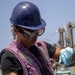 A worker wearing a blue hard hat, safety goggles, blue nitrile gloves, and a purple vest is handling a clear glass bottle containing a green/yellow liquid. The setting appears to be an outdoor industrial or maritime environment, with what looks like a naval vessel or industrial structure visible in the background. The activity suggests fuel or liquid sampling, consistent with maritime or military fueling operations.
