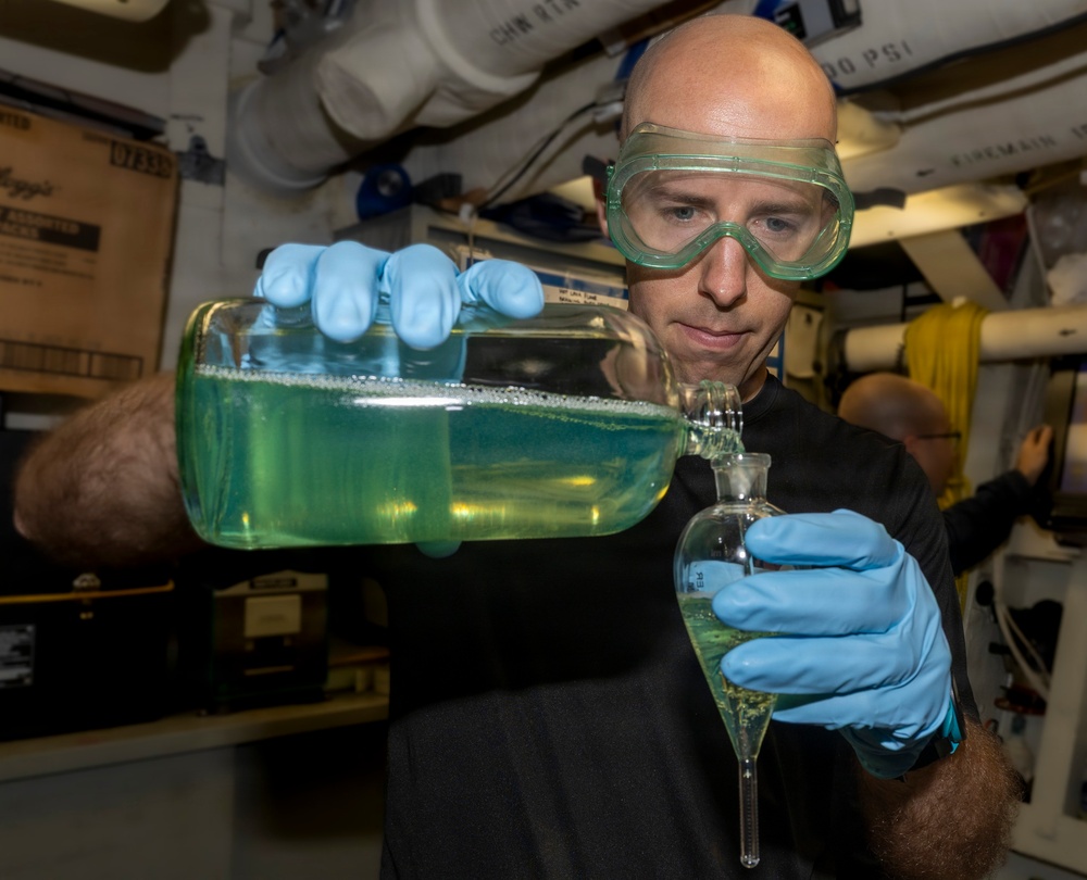 A bald man wearing green safety goggles and blue nitrile gloves is carefully pouring a green liquid from a large rectangular glass container into a separatory funnel. The activity appears to be taking place in a confined, industrial or shipboard laboratory environment. The careful, focused demeanor and protective equipment suggest a controlled chemical or water-testing procedure.