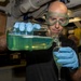 A bald man wearing green safety goggles and blue nitrile gloves is carefully pouring a green liquid from a large rectangular glass container into a separatory funnel. The activity appears to be taking place in a confined, industrial or shipboard laboratory environment. The careful, focused demeanor and protective equipment suggest a controlled chemical or water-testing procedure.