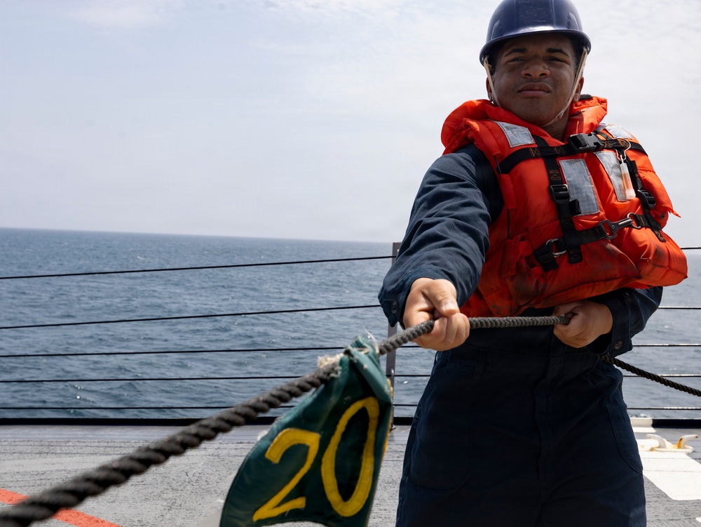 A sailor or naval personnel is working on the deck of a ship at sea, pulling a thick rope while wearing standard maritime safety equipment. The individual is positioned near a green marker labeled '20', likely a deck position or mooring marker. The ocean is visible in the background under an overcast sky.