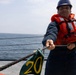 A sailor or naval personnel is working on the deck of a ship at sea, pulling a thick rope while wearing standard maritime safety equipment. The individual is positioned near a green marker labeled '20', likely a deck position or mooring marker. The ocean is visible in the background under an overcast sky.