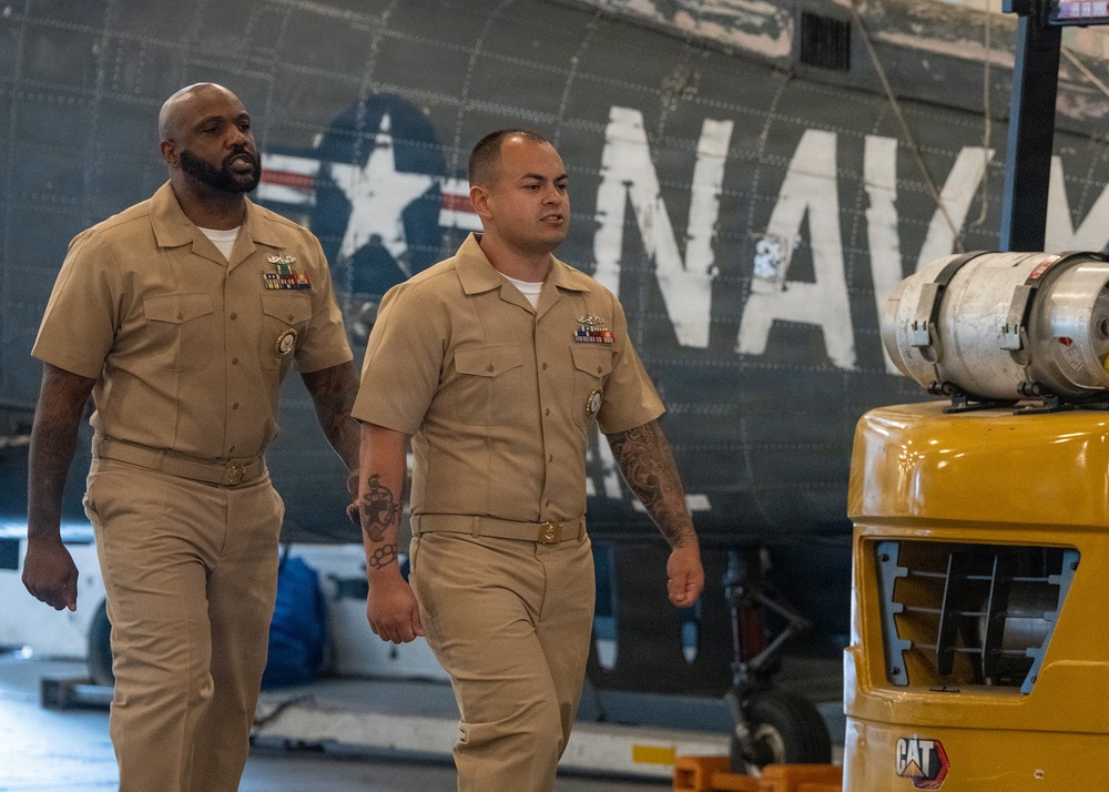 Two U.S. Navy enlisted personnel in Service Khaki uniforms walk through what appears to be a hangar bay or aviation maintenance facility. Both sailors display ribbons and warfare/qualification badges on their uniforms, indicating experienced service members. A vintage Navy aircraft with 'NAVY' markings and a star insignia is visible in the background, along with a yellow CAT ground support vehicle.