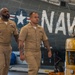 Two U.S. Navy enlisted personnel in Service Khaki uniforms walk through what appears to be a hangar bay or aviation maintenance facility. Both sailors display ribbons and warfare/qualification badges on their uniforms, indicating experienced service members. A vintage Navy aircraft with 'NAVY' markings and a star insignia is visible in the background, along with a yellow CAT ground support vehicle.