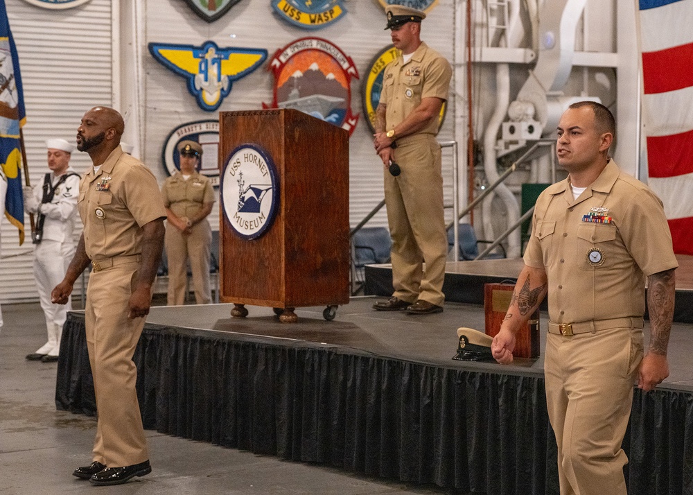 A formal U.S. Navy ceremony is taking place at the USS Hornet Museum, as indicated by the podium bearing the museum's logo. Multiple Navy personnel in tan service uniforms (Service Khakis) are present on and around a draped stage, with a color guard member in dress whites visible in the background. The setting is decorated with various naval unit patches and insignia on the walls.