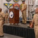 A formal U.S. Navy ceremony is taking place at the USS Hornet Museum, as indicated by the podium bearing the museum's logo. Multiple Navy personnel in tan service uniforms (Service Khakis) are present on and around a draped stage, with a color guard member in dress whites visible in the background. The setting is decorated with various naval unit patches and insignia on the walls.