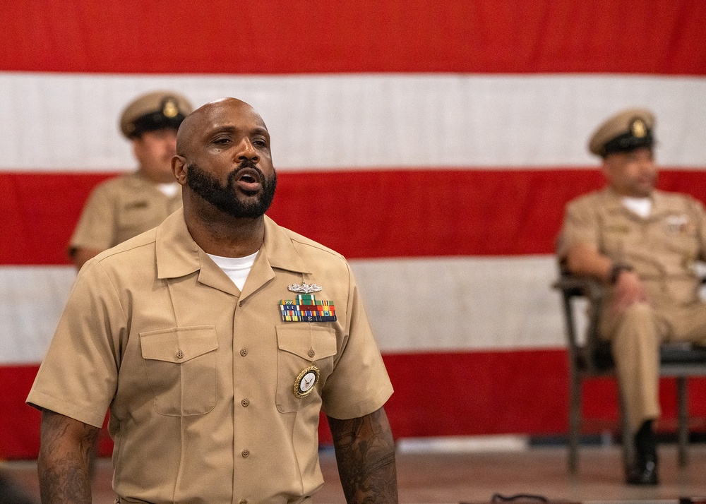 A U.S. Navy enlisted sailor in khaki service uniform stands in the foreground, appearing to be speaking or reciting something at a formal military ceremony. Two other Navy personnel in similar uniforms are seated in the background. A large American flag with red and white stripes serves as a backdrop.