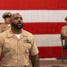 A U.S. Navy enlisted sailor in khaki service uniform stands in the foreground, appearing to be speaking or reciting something at a formal military ceremony. Two other Navy personnel in similar uniforms are seated in the background. A large American flag with red and white stripes serves as a backdrop.