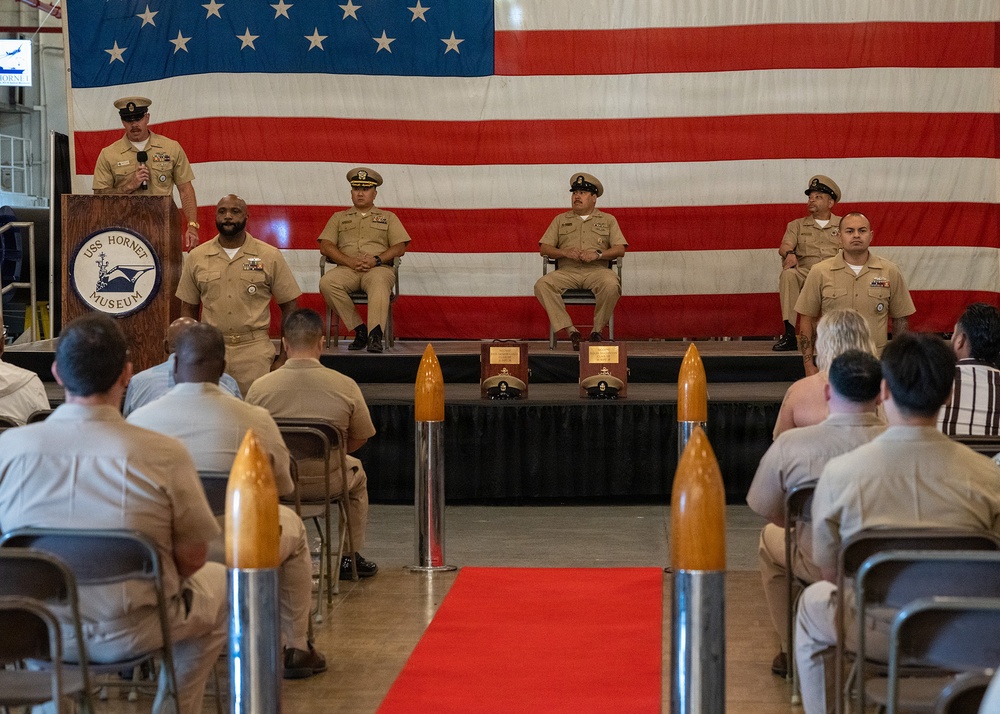 A formal U.S. Navy ceremony is taking place aboard or at the USS Hornet Museum, with personnel in khaki service uniforms gathered in an auditorium-style setting. An officer is speaking at a podium bearing the USS Hornet Museum seal, while senior enlisted or officer personnel are seated on a raised stage behind him. A large American flag serves as a backdrop, and decorative shell casings flank a red carpet leading to the stage.