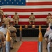 A formal U.S. Navy ceremony is taking place aboard or at the USS Hornet Museum, with personnel in khaki service uniforms gathered in an auditorium-style setting. An officer is speaking at a podium bearing the USS Hornet Museum seal, while senior enlisted or officer personnel are seated on a raised stage behind him. A large American flag serves as a backdrop, and decorative shell casings flank a red carpet leading to the stage.