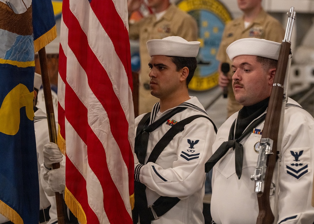 A U.S. Navy color guard is participating in a formal military ceremony, with sailors in dress white uniforms carrying the American flag and a rifle. At least two petty officers are prominently visible in the foreground, wearing traditional Navy dress whites with black neckerchiefs and service ribbons. The background shows additional Navy personnel in khaki uniforms near what appears to be a unit insignia or patch on a wall.