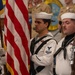 A U.S. Navy color guard is participating in a formal military ceremony, with sailors in dress white uniforms carrying the American flag and a rifle. At least two petty officers are prominently visible in the foreground, wearing traditional Navy dress whites with black neckerchiefs and service ribbons. The background shows additional Navy personnel in khaki uniforms near what appears to be a unit insignia or patch on a wall.